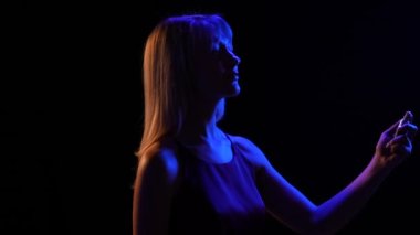 Üzerine sprey şişesinden parfüm serpiştiren genç ve güzel bir sarışın siluetinin yan görüntüsü. Siyah stüdyo arka planında mavi neon ışıklarla poz veren güzel bir kızın portresi. Kapatın. Yavaş çekim