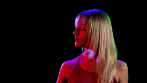 A young pretty blonde sprinkling perfume on herself from a spray bottle and enjoying its scent. Portrait of a cute girl posing over black studio background with neon lights. Close up. Slow motion. — Stock Video