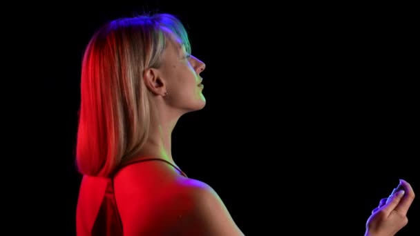 Side view of young pretty blonde sprinkling perfume on herself from a spray bottle. Portrait of a cute girl posing over black studio background with neon lights. Close up. Slow motion. — Stock Video