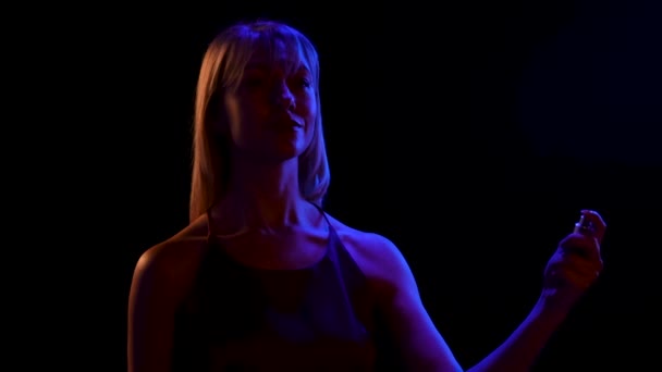 Silhouette of a young pretty blonde sprinkling perfume on herself from a spray bottle. Portrait of a cute girl posing over black studio background with blue neon lights. Close up. Slow motion. — Stock Video