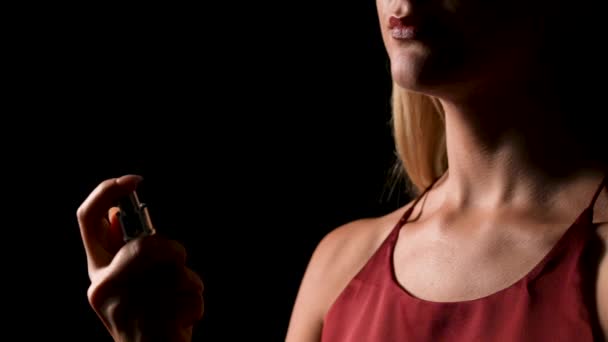 A young pretty blonde sprinkling perfume on herself from spray bottle. Portrait of cute girl posing on black studio background with backlight. Part of face is hidden in shadow. Close up. Slow motion. — Stock Video
