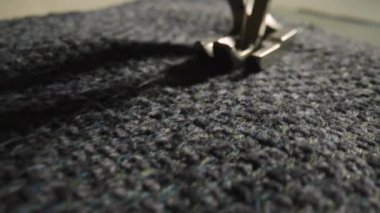 Movement of foot and needle while sewing gray woolen fabric with sewing machine. Silver presser foot of the sewing machine presses the fabric down and sews it with a needle. Close up. Slow motion.