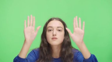 Portrait of an excited young girl banging her fists on glass, waving, calling someone and gesturing I am here. Model girl posing in studio against background of green screen. Close up. Slow motion.