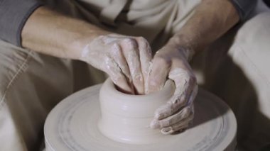 Çömlekçi erkek elleri, yaratıcı bir atölyede çömlekçi çarkının üzerinde dönen yumuşak beyaz kil tenceresini şekillendirir ve şekillendirir. Usta çömlekçiliğe farklı şekil verir. Clay yakından şekilleniyor. Yavaş çekim.