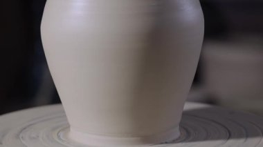 Yaratıcı bir atölyede çömlekçi çarkında dönen yumuşak, beyaz bir çömlek parçası. Çömlekçi tarafından şekillendirilmiş bir çömlek. Toprak kapları yumuşak ışıkta kapanır. Yavaş çekim.