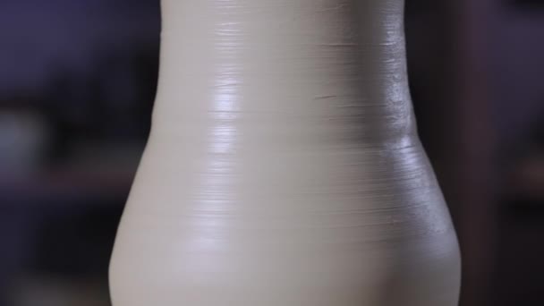 Een vormgegeven stuk zacht wit aardewerk, draaiend op een pottenbakkerswiel in een creatieve werkplaats. Een pot gevormd door de handen van een pottenbakker. Aardewerk sluiten in zacht licht. Langzame beweging. — Stockvideo