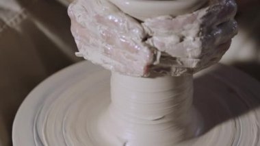 Çömlekçi erkek elleri, yaratıcı bir atölyede çömlekçi çarkının üzerinde dönen yumuşak beyaz kil tenceresini şekillendirir ve şekillendirir. Usta çömlekçiliğe şekil verir. Clay yakından şekilleniyor. Yavaş çekim.