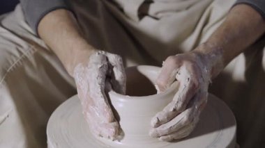 Çömlekçi erkek elleri, yaratıcı bir atölyede çömlekçi çarkının üzerinde dönen yumuşak beyaz kil tenceresini şekillendirir ve şekillendirir. Usta çömlekçiliğe şekil verir. Clay yakından şekilleniyor. Yavaş çekim.