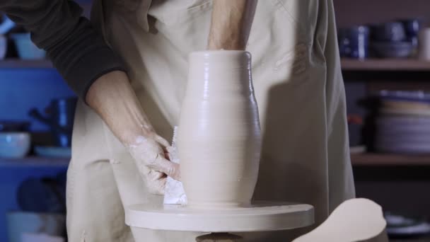 Man handen van pottenbakker vormen een zachte witte klei pot met behulp van aardewerk gereedschap, draaien het op een pottenbakkerswiel in een creatieve werkplaats. De meester geeft vorm aan het aardewerk. Clay maakt een close-up. Langzame beweging. — Stockvideo