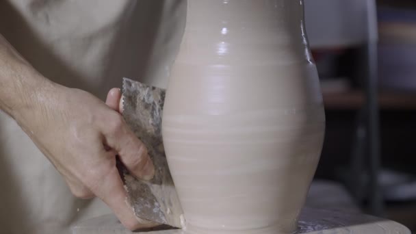 Male hands of potter form a soft white clay pot using pottery tools, turning it on a potters wheel in a creative workshop. The master gives a shape to the pottery. Clay shaping close up. Slow motion. — Stock Video