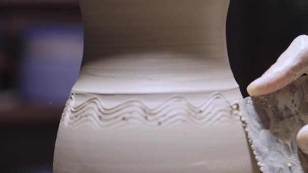 De manliga händerna på en krukmakare snida mönster på en mjuk vit lerkruka med hjälp av keramik verktyg. Keramiska varor roterar på ett keramikhjul i en kreativ verkstad. Earthenware närbild i mjukt ljus. Långsamma rörelser. — Stockvideo