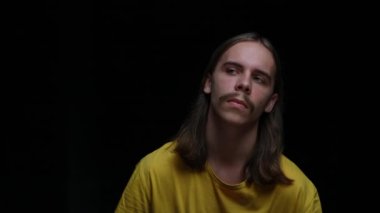 Genç bir adamın portresi ilgi çekici bir şekilde kameraya bakıyor ve bana bir jest yapıyor. Stüdyoda siyah arka planda poz veren uzun saçlı ve bıyıklı bir erkek model. Kapatın. Yavaş çekim.