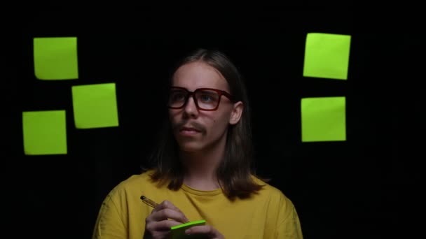 Retrato del joven escribiendo sus pensamientos con bolígrafo en notas adhesivas, pegándolas en vidrio y regocijándose ante una buena idea. Modelo masculino posando sobre fondo negro en estudio. De cerca. Movimiento lento. — Vídeos de Stock