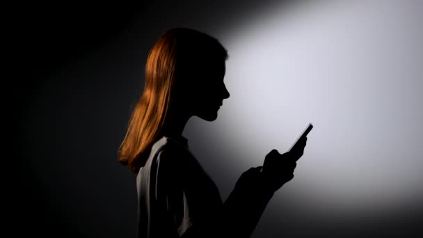 Sidovy siluett av en ung flicka sms:a på sin telefon och vara mycket glad. Flickmodell poserar i mörk studio med iscensatt framljus. Närbild. Långsamma rörelser. — Stockvideo