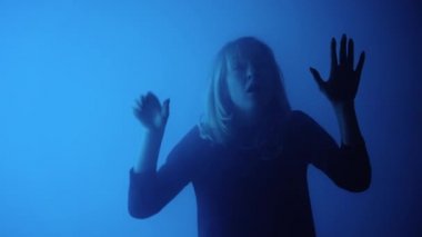 Kadın dumandan çıkar ve korkunç bir şekilde cama avuç içiyle dokunur. Sarışın, elleri cama kayar, korku ve dehşetle bağırır. Model, karanlık stüdyoda mavi ışıkların arka planına karşı poz veriyor. Yakın çekim.