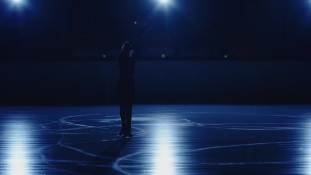 Joven patinadora femenina realiza coreografía de patinaje individual en pista de hielo sobre fondo de suave luz azul. Silueta de un atleta deslizándose sobre hielo practicando saltos y giros. Movimiento lento. — Vídeos de Stock