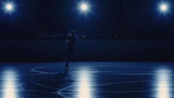 Samice umělecké krasobruslařka provádí jednu bruslení choreografii na kluzišti proti měkkému modrému světlu. Silueta sportovce provádějícího rotaci na jedné noze. Zpomalení otáčení v krasobruslení — Stock video