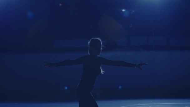 Jonge vrouwelijke kunstschaatsster voert enkele schaatschoreografie uit op de ijsbaan tegen zacht blauw licht. Silhouet van een atleet glijden op ijs oefenen sprongen en spins. Langzame beweging. Sluiten.. — Stockvideo