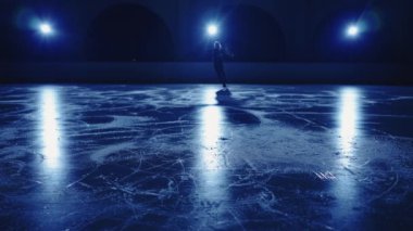 Genç bayan artistik patinajcının bulanık silueti, yumuşak mavi ışığa karşı buz arenasında tek bir paten koreografisi sergiliyor. Atlet kadın kayma ve dönme becerileri üzerinde çalışıyor. Yavaş çekim.