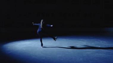 Genç artistik patinajcı kız mavi bir spot ışığı altında buz arenasında zarif bir şekilde kayıyor. Tam boy atış, profesyonel artistik patinajcı tek bacakla antrenman yapıyor. Yavaş çekim.