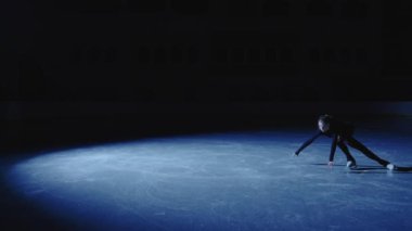 Genç artistik patinajcı kız mavi bir spot ışığı altında buz arenasında zarif bir şekilde kayıyor. Tam boy atış, profesyonel artistik patinajcı tek bacakla antrenman yapıyor. Yavaş çekim.
