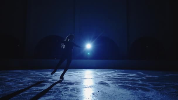 Размытый силуэт фигуристки катается по ледяной арене в темноте, женская фигура движется против синей софиты в замедленной съемке. Полнометражный снимок, практикующий навыки скольжения и вращения. — стоковое видео