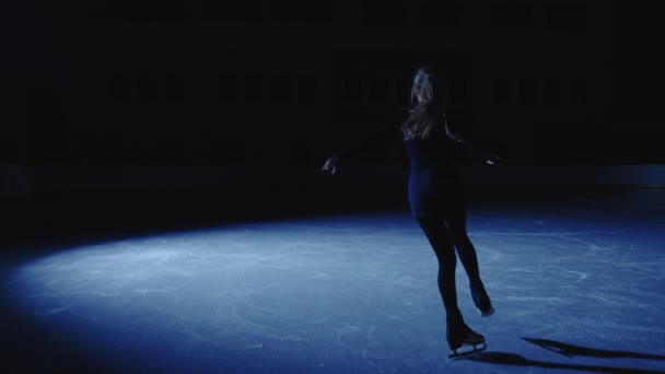Νεαρό κορίτσι πατινάζ φιγούρα χαριτωμένα διαφάνειες στον πάγο στην αρένα πάγου υπό το φως ενός μπλε προβολέα. Full length shot, επαγγελματική φιγούρα skater εξάσκηση περιστροφή σε μια εσωτερική αρένα πάγου. Αργή κίνηση. — Αρχείο Βίντεο