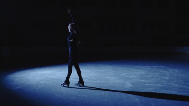 Vrouw verschijnt op het ijs arena in blauw licht van schijnwerpers en glijdt over ijs, het beoefenen van enkele choreografie. Vrouwelijke kunstschaatser bereidt zich voor op prestaties en training rotatie vaardigheden. Langzame beweging. — Stockvideo