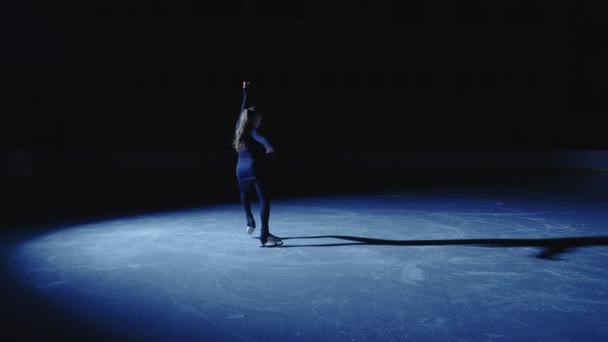 Jonge kunstschaatsster schuift sierlijk op het ijs in de ijshal in het licht van een blauwe schijnwerper. Volledige shot, professionele kunstschaatser oefenen spin op een been. Langzame beweging. — Stockvideo