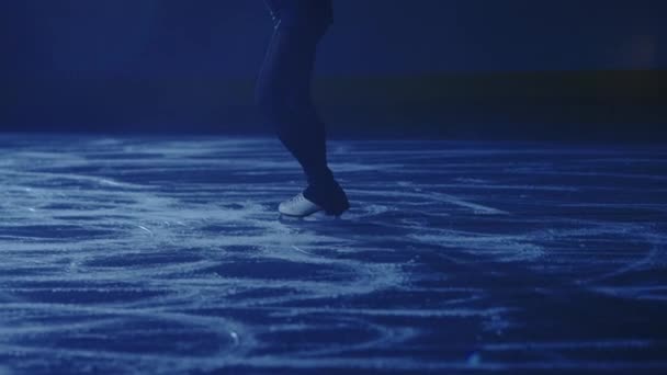 Фігурна дама катається на ковзанах, тренуючись наодинці вночі в променях блакитного світла. Молода жінка готується до змагань, тренує навички ротації. Повільний рух, ноги крупним планом . — стокове відео