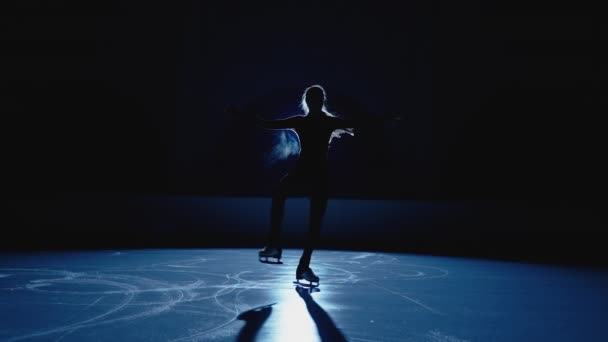 Junge Eiskunstläuferin rotiert in dunkler Eisarena vor hellblauem Scheinwerferlicht. Silhouette einer Athletin, die Drehungen für eine Eisshow übt. Eine einzige Choreographie. Zeitlupe. — Stockvideo