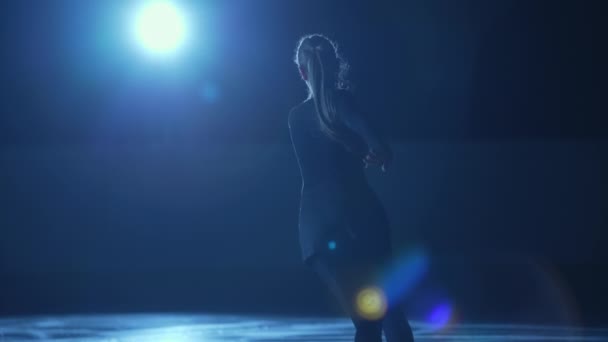 Junge Eiskunstläuferin rotiert in dunkler Eisarena vor hellblauem Scheinwerferlicht. Silhouette einer Athletin, die Drehungen für eine Eisshow übt. Eine einzige Choreographie. Zeitlupe. — Stockvideo