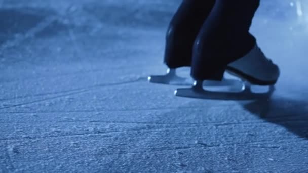 Detalle de tiro de piernas de mujer en patines de patinaje artístico deslizándose sobre hielo frío en arena. Una deportista profesional entrena en la oscuridad con luz azul. La mujer se desliza sobre el hielo y se detiene. De cerca. Movimiento lento. — Vídeos de Stock