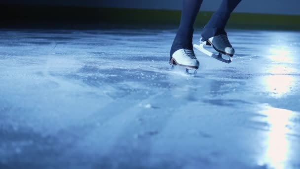 Detailní záběr ženských nohou v krasobruslích klouzajících na ledě v aréně ve tmě s modrým světlem. Žena sklouzne po ledu a rozstříkne do kamery částečky třpytivého ledu. Zavřít. Zpomalený pohyb. — Stock video