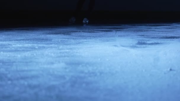 Foto detallada de las piernas de las mujeres en patines de patinaje artístico deslizándose sobre hielo en una arena en la oscuridad con luz azul. La mujer se desliza sobre el hielo, salpicando partículas de hielo espumoso en la cámara. De cerca. Movimiento lento. — Vídeos de Stock