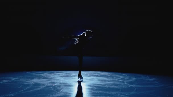 어린 피겨 스케이팅 아티스트는 밝은 푸른 조명을 배경으로 얼음 경기장에서 어두운 곳에서 회전한다. 아이스 쇼를 위해 스핀을 연습하는 여성 운동 선수 실루엣. 단수 안무. 느린 동작. — 비디오