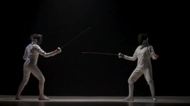 Siyah arka planda bir rapier ile kadın eskrimcilerin eğitimi. Beyaz üniformalı ve eskrim maskeli sporcular bu beceriyi geliştiriyor, düşmana saldırıyor, birbirlerine vuruyorlar, rekabetçi bir düello. Yavaş çekim.