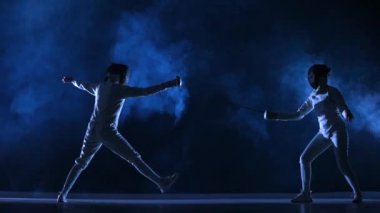 İki bayan sporcu eskrim turnuvasında folyo üzerinde antrenman yapıyor. Beyaz üniformalı ve eskrim maskeli genç silahşörler dumanlı ve mavi ışıkla karanlık stüdyoda düello yapıyorlar. Eskrim okulu. Yavaş çekim