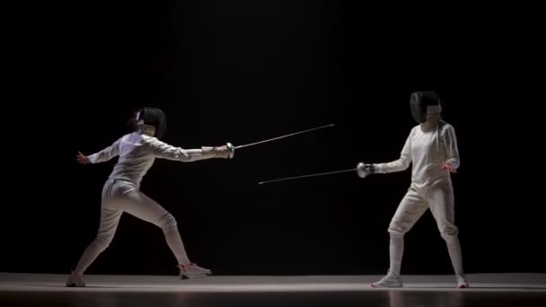 2人の女性選手がフェンシングトーナメントでホイルで訓練する。白い制服を着た若い女性剣士とフェンシングマスクは黒を背景に決闘を戦う。フェンシングのスポーツスクール。スローモーション. — ストック動画