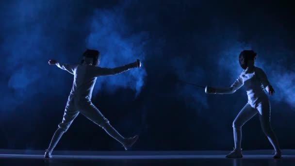 Two female athletes train on foil at fencing tournament. Young swordsmen in white uniforms and fencing masks fight duel on dark studio background with smoke and blue light. Fencing school. Slow motion — Stock Video