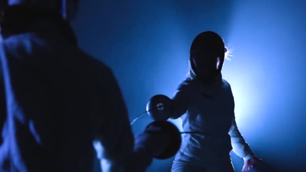 Вид сзади на двух спортсменов-фехтовальщиков, соревнующихся в турнире. Женщины в защитных масках нападают друг на друга, ударяя эпинефрином. Темная студия с подсветкой синим светом и дымом. Медленное движение. Закрыть. — стоковое видео
