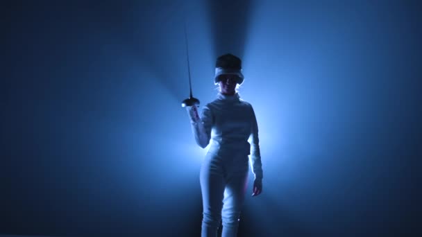 Портрет молодой женщины-фехтовальщицы, надевающей маску и занимающей боевую позицию с мечом в руке. Спортсмен позирует в темной студии с подсветкой синего света и дыма. Медленное движение. Закрыть. — стоковое видео