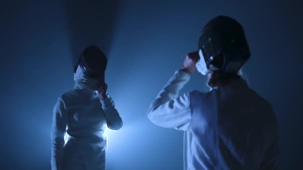 Two young female fencers take off their fencing masks salute with rapiers and greet each other. Womans hitting palm on palm in dark studio with backlit blue light and smoke. Slow motion. Close up. — Stock Video