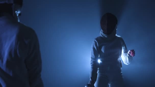 View from behind on two athletes fencers competing in tournament. Women in fencing masks attack each other, striking with epee. Dark studio with backlit blue light and smoke. Slow motion. Close up. — Stock Video