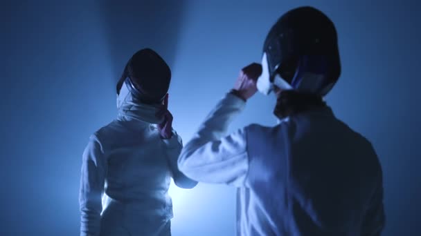 Дві молоді жінки-фехтувальники знімають маски для фехтування і вітають один одного. Жінки б'ють долонею на долоні в темній студії з підсвічуванням синім світлом і димом. Повільний рух. крупним планом . — стокове відео