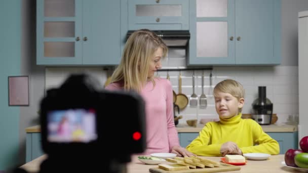 Young woman teaches boy how to make sandwiches and filming video with camera. Mom and son filming stories in kitchen for social media cooking blog. Family bloggers. Slow motion ready, 4K at 59.97fps. — Stock Video