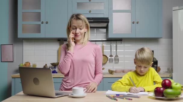 A nő laptopon dolgozik, telefonon beszél, a fiú filctollal rajzol. Anya és fia a konyhában. Családi bloggerek történeteket filmeznek a közösségi médiának és az élet videó blognak. Lassított felvétel kész, 59.97fps.. — Stock videók