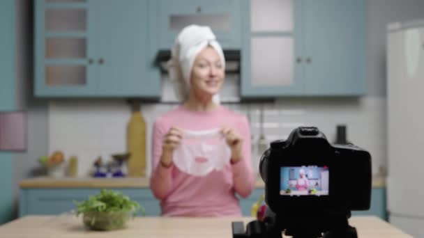 Jovem fala com a câmera sobre os benefícios do uso de máscara facial de folha hidratante. Blogueira feminina filmando histórias na cozinha para blog de beleza de mídia social. Movimento lento pronto, 4K a 59.97fps. — Vídeo de Stock