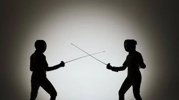 決闘を戦う2人の女性剣士の黒いシルエット。選手はレイピアを渡り、攻撃的な打撃を提供します。スポットライトのある白い背景の暗いスタジオに女の子たちがポーズをとっている。スローモーション. — ストック写真
