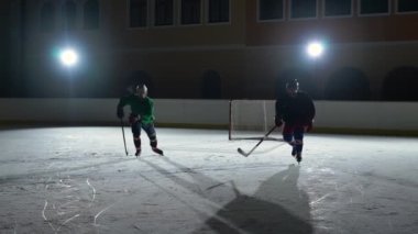 Üniformalı ve kasklı iki adam buz pateni sahasında kayıyor ve birbirleriyle çarpışıyorlar. Profesyonel hokey oyuncuları spot ışıklarıyla karanlık bir pistte antrenman yaparlar. Hokey Spor Okulu. Yavaş çekim.
